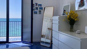 A room with a large sliding glass door leading to a balcony with a view of the sea. There is a white dresser with a mirror and a vase of yellow flowers on top.