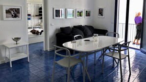 A modern living room with a black sofa, a white dining table, and a glass coffee table. There are framed pictures on the wall and a balcony with a person outside.