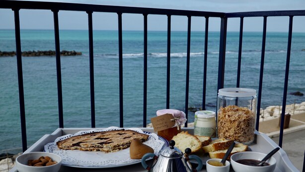 A tray with breakfast items including bread, a pie, and a jar of cereal, set against a seaside view.