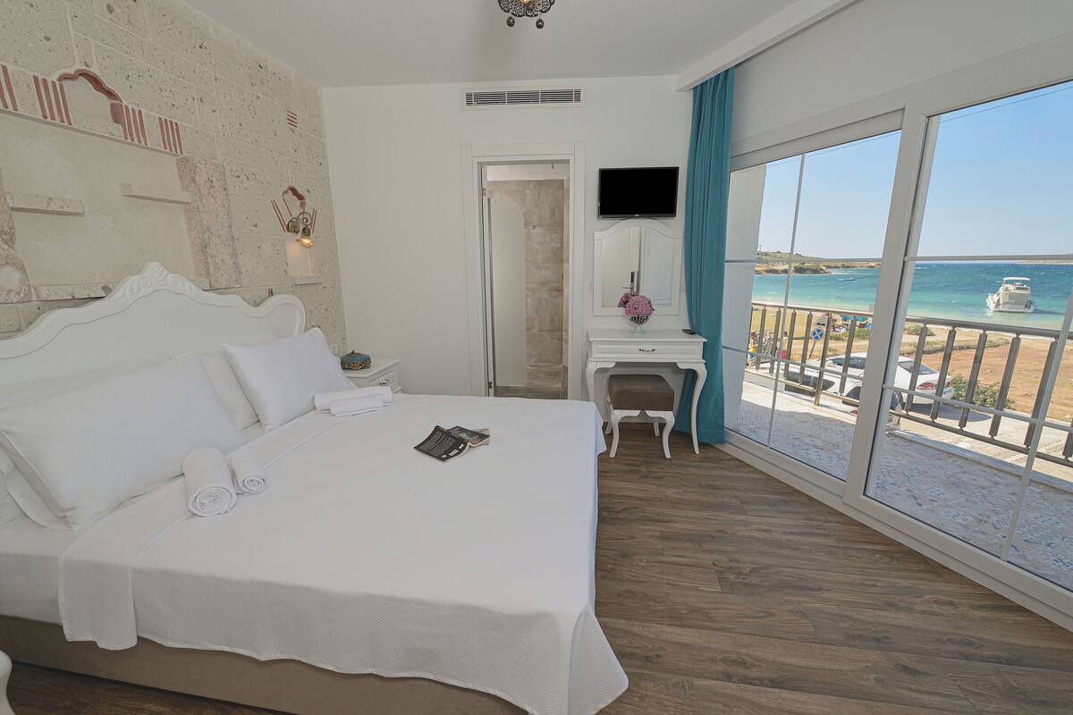 A hotel room with a large bed, a desk, a TV, and a balcony with a view of the sea.