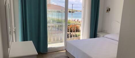 Un dormitorio con una cama grande, una mesita de noche, un aparador y vistas a una playa y al mar a través de un ventanal.
