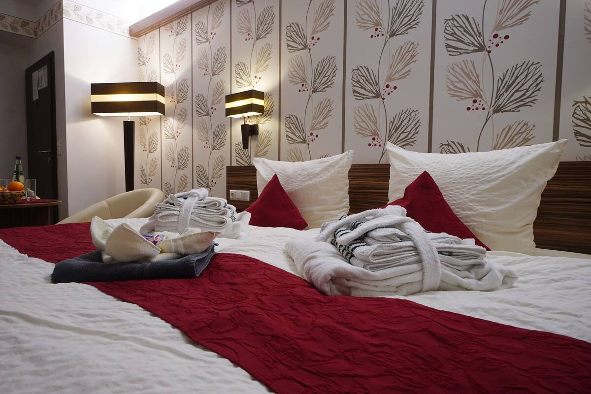 A neatly made bed with towels, a red throw blanket, and pillows. There is a bedside table with a fruit bowl and glasses. The room has floral wallpaper and a modern lamp.