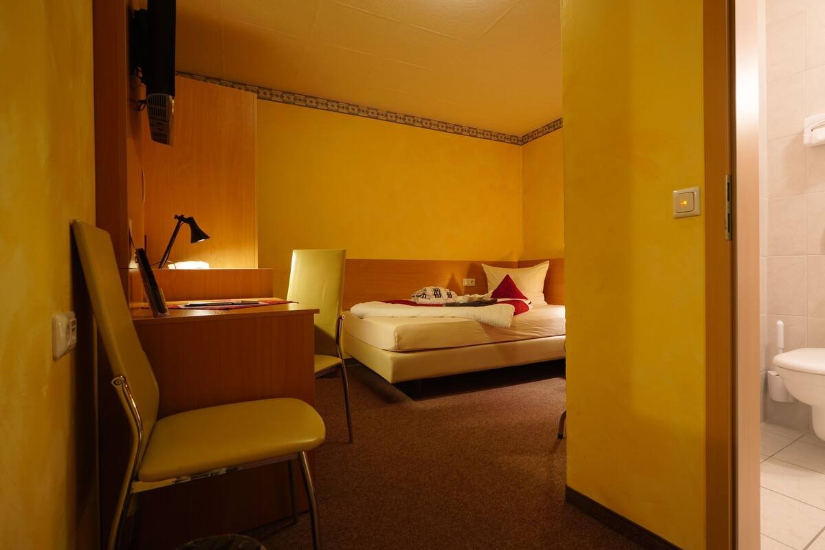 A hotel room with a bed, a desk with a lamp, a chair, and a bathroom with a toilet and towel rack.