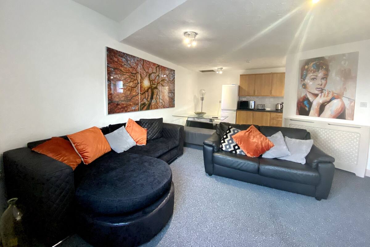 Ein modernes Wohnzimmer mit zwei schwarzen Sofas, einem Couchtisch und einem großen abstrakten Gemälde. Im Hintergrund ist eine Küche mit Holzschränken und einer Mikrowelle zu sehen.
