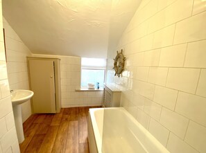 A narrow bathroom with a wooden floor, a white bathtub, a sink, and a window with a view. - Simpson Beach View Apartments (Blackpool)