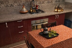 A kitchen with a Moroccan tagine, a coffee pot, and teapots on the counter, a stove, and a table with a floral tablecloth. - 🔥 Appartement à Mehdya - Plage à proximité - Idéal famille (kènitra)
