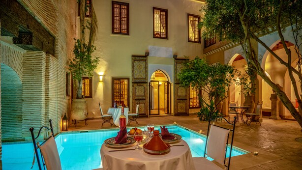 A poolside dining area with a table set for two, surrounded by chairs, and a pool in the background. - Il Parco dei Principi (Marrakech)
