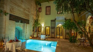 An indoor swimming pool with a tiled floor, surrounded by a patio area with chairs and tables, and a brick wall with windows. - Il Parco dei Principi (Marrakech)
