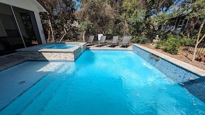 Une piscine rectangulaire dont le bord est recouvert de carreaux, entourée d’une bordure en pierre et d’une clôture en métal. Des chaises longues sont disposées le long du bord de la piscine.