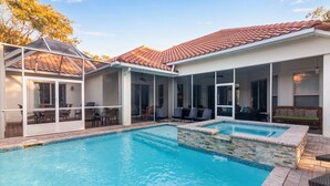 Una piscina con solárium de baldosas, un patio cubierto con sillas y un edificio con techo de tejas.