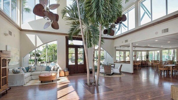 Un salón espacioso con piso de madera, un área de estar central con sofá y reposapiés, una palmera alta y un comedor con mobiliario de madera.