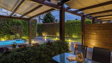 Een terras buiten met een glazen tafel, stoelen, een jacuzzi en een houten hek.