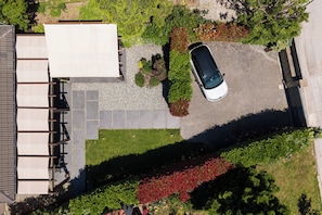 Luchtbeeld van een woongebied met een witte auto op een oprit, een huis met een pannendak en een tuin met verschillende planten en struiken.