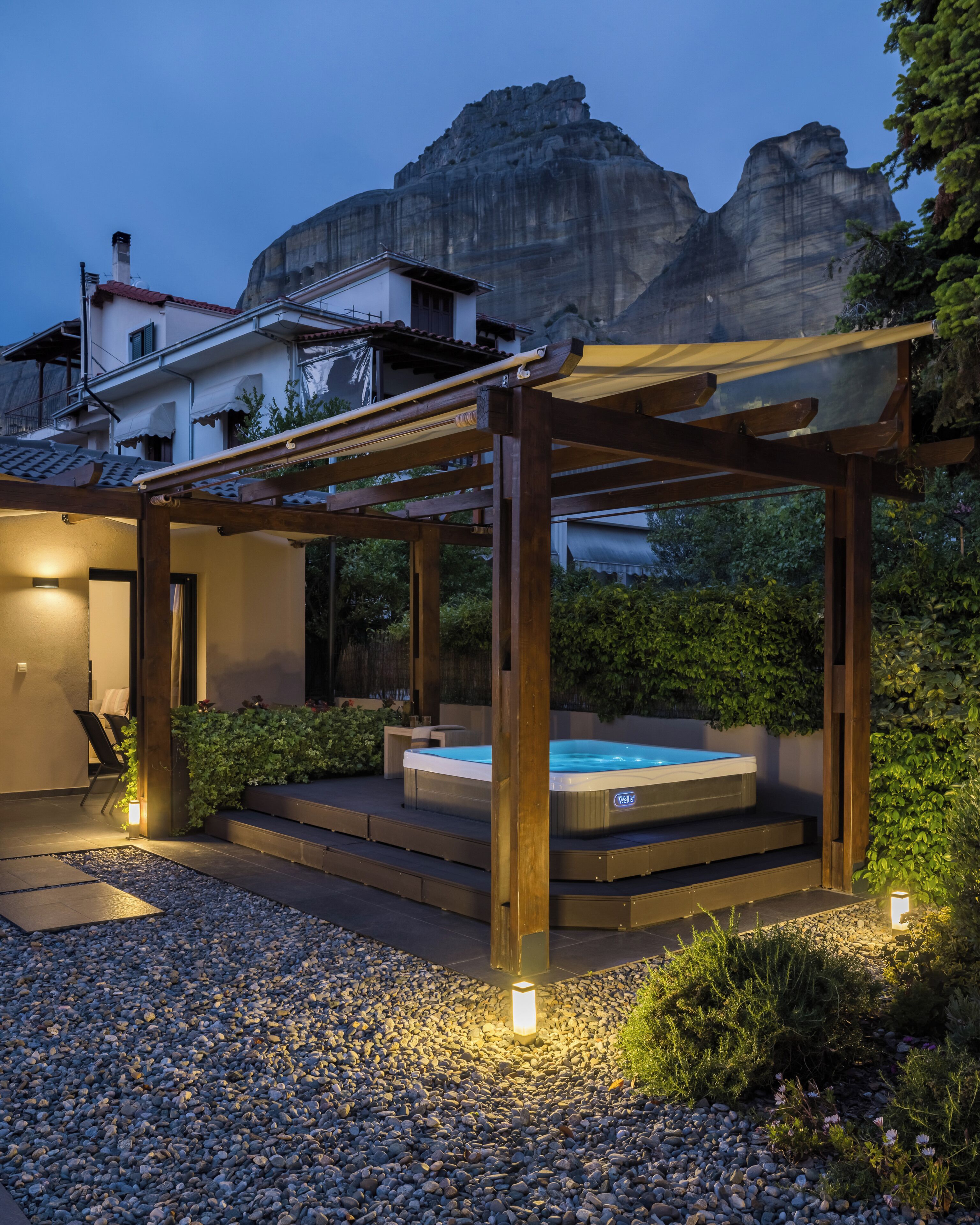 A hot tub under a wooden pergola with steps leading up to it, surrounded by gravel and plants.