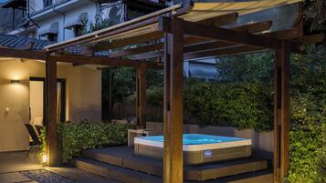 A hot tub under a wooden pergola with steps leading up to it, surrounded by gravel and plants.