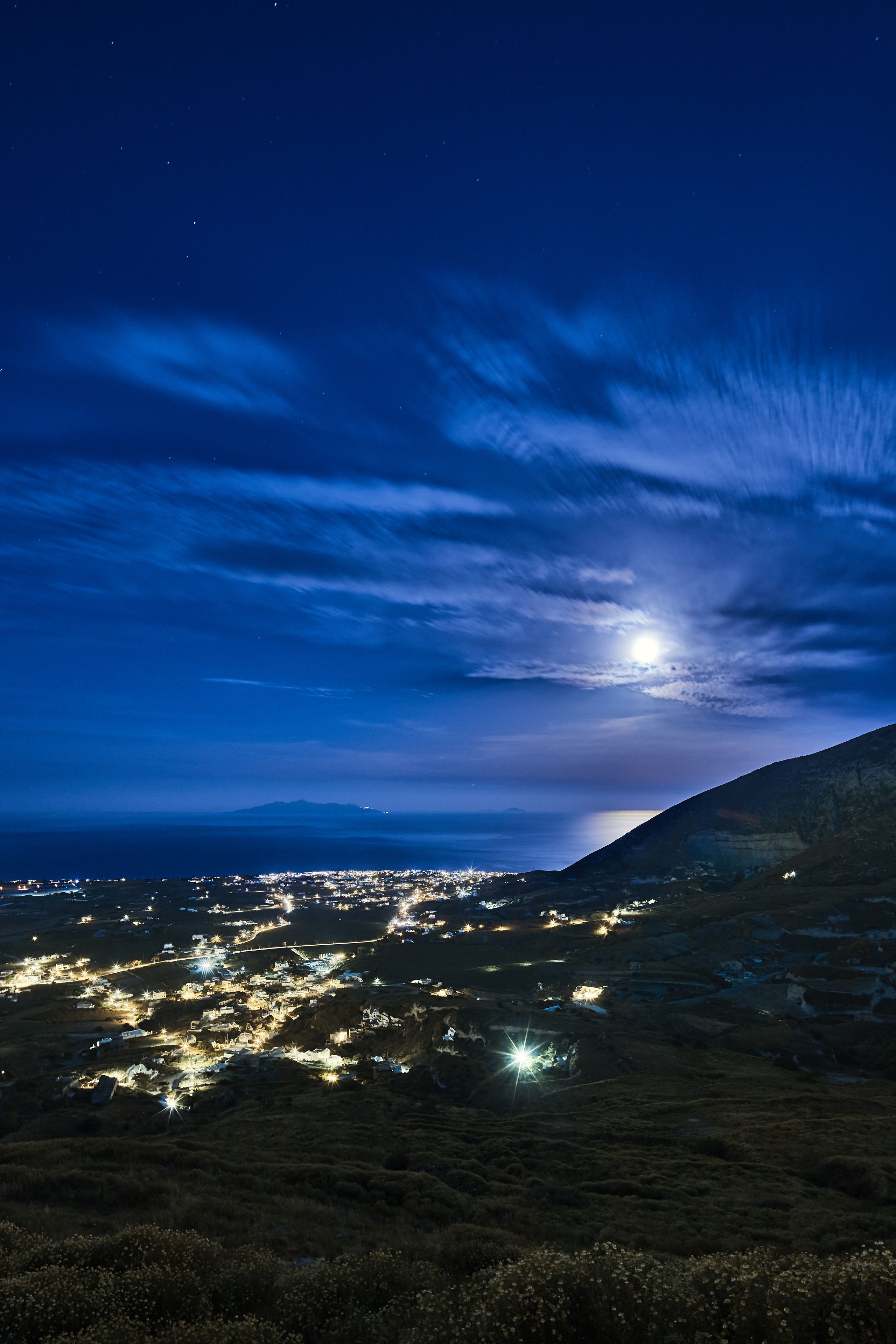 A night view of a coastal town with a prominent hill and a visible moon.