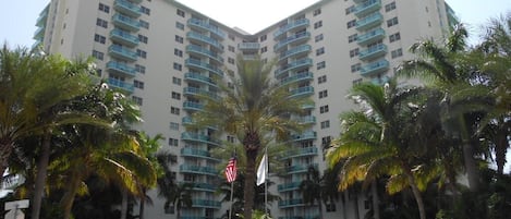 Un hotel de varios pisos con un patio central, palmeras y un asta con bandera.