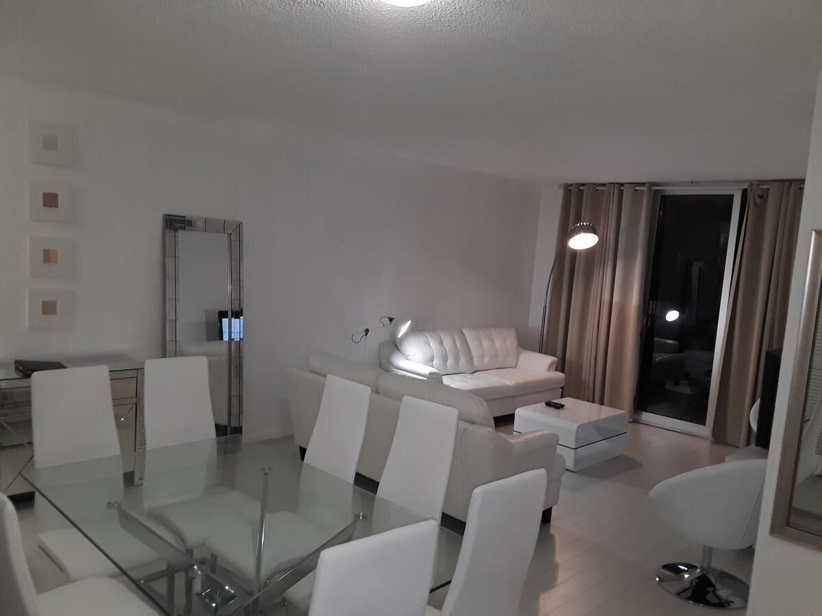 A modern living room with a glass dining table, white sofas, and a sliding glass door leading to a balcony.