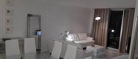A modern living room with a glass dining table, white sofas, and a sliding glass door leading to a balcony.