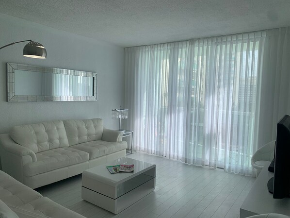 Un salón moderno con un sofá blanco, una mesa de centro de vidrio, una lámpara de pie y cortinas translúcidas.