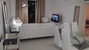 Ein modernes Wohnzimmer mit einem weißen Sofa, einem Glastisch, einem Flachbildfernseher und einem großen Fenster mit Vorhängen.