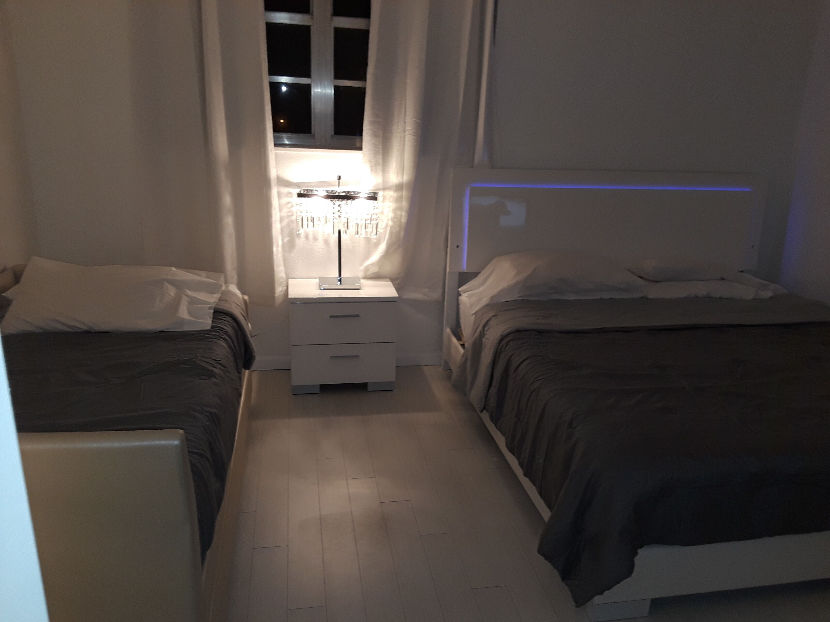 Habitación de hotel estrecha con dos camas, una mesita de noche con lámpara y una ventana con cortinas.