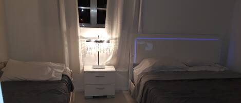 Habitación de hotel estrecha con dos camas, una mesita de noche con lámpara y una ventana con cortinas.