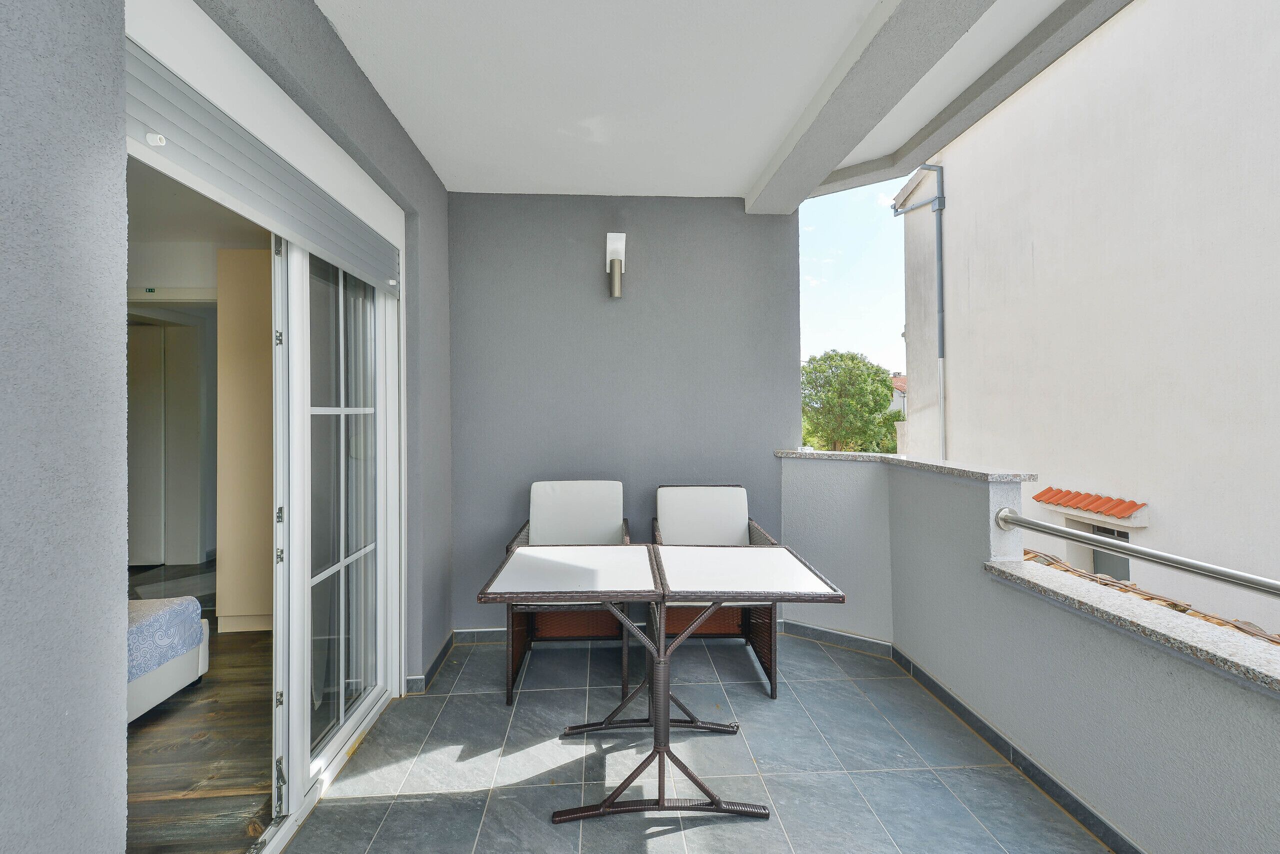 A balcony with a table and two chairs, a sliding glass door, and a view of a tree.