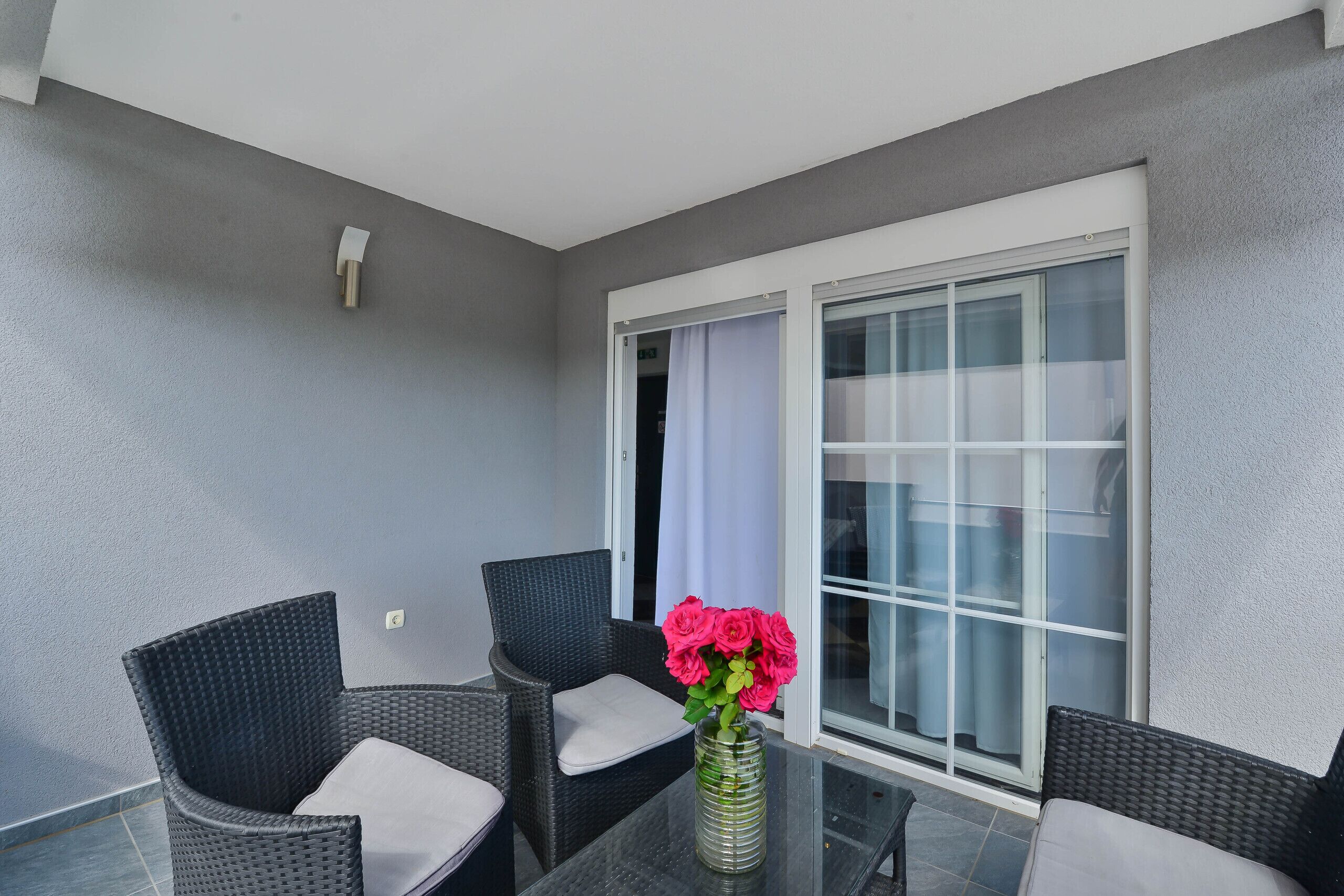 Un balcone con due sedie in vimini, un tavolino in vetro e un vaso con fiori.