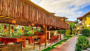 Uma área de piscina com mesas e cadeiras de madeira, um espaço coberto para relaxar e um caminho que leva a um edifício com fachada amarela.