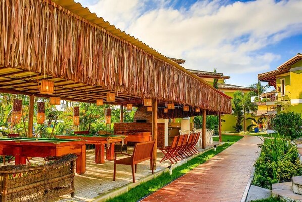 A pool area with wooden tables and chairs, a covered seating area, and a pathway leading to a building with a yellow facade.