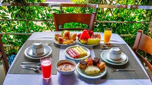 Ein gedeckter Tisch mit Frühstücksleckereien, darunter Früchte, Gebäck und Getränke, umgeben von üppigem Grün.