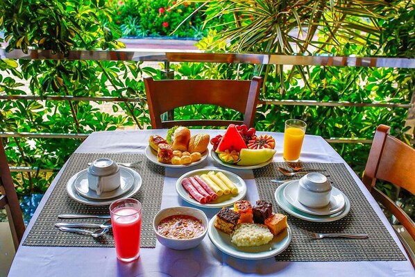 Une table dressée avec des aliments pour le petit-déjeuner, notamment des fruits, des viennoiseries et des boissons, entourée d’une végétation luxuriante.