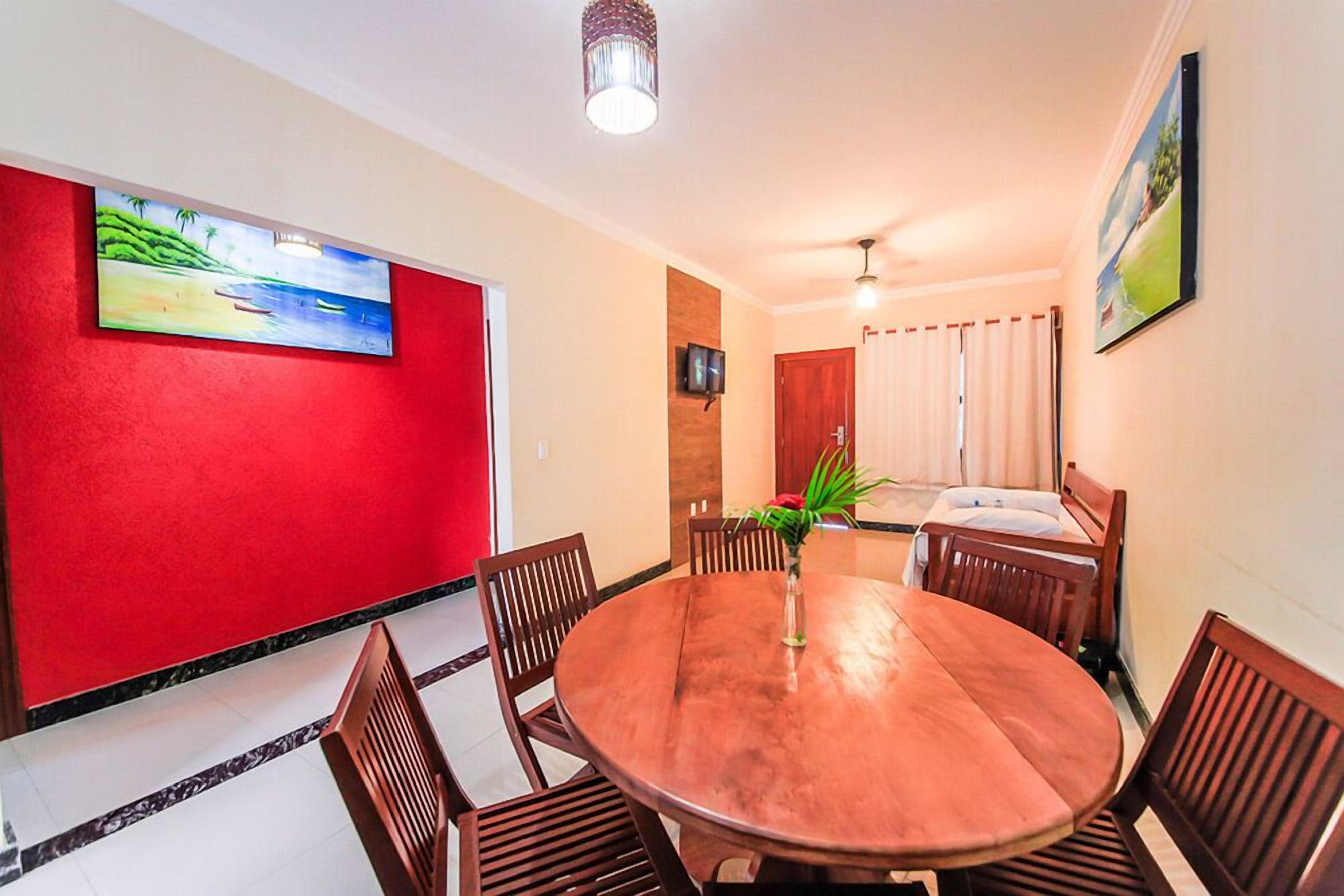 A dining area with a round wooden table and chairs, a red accent wall, and a painting above the table.