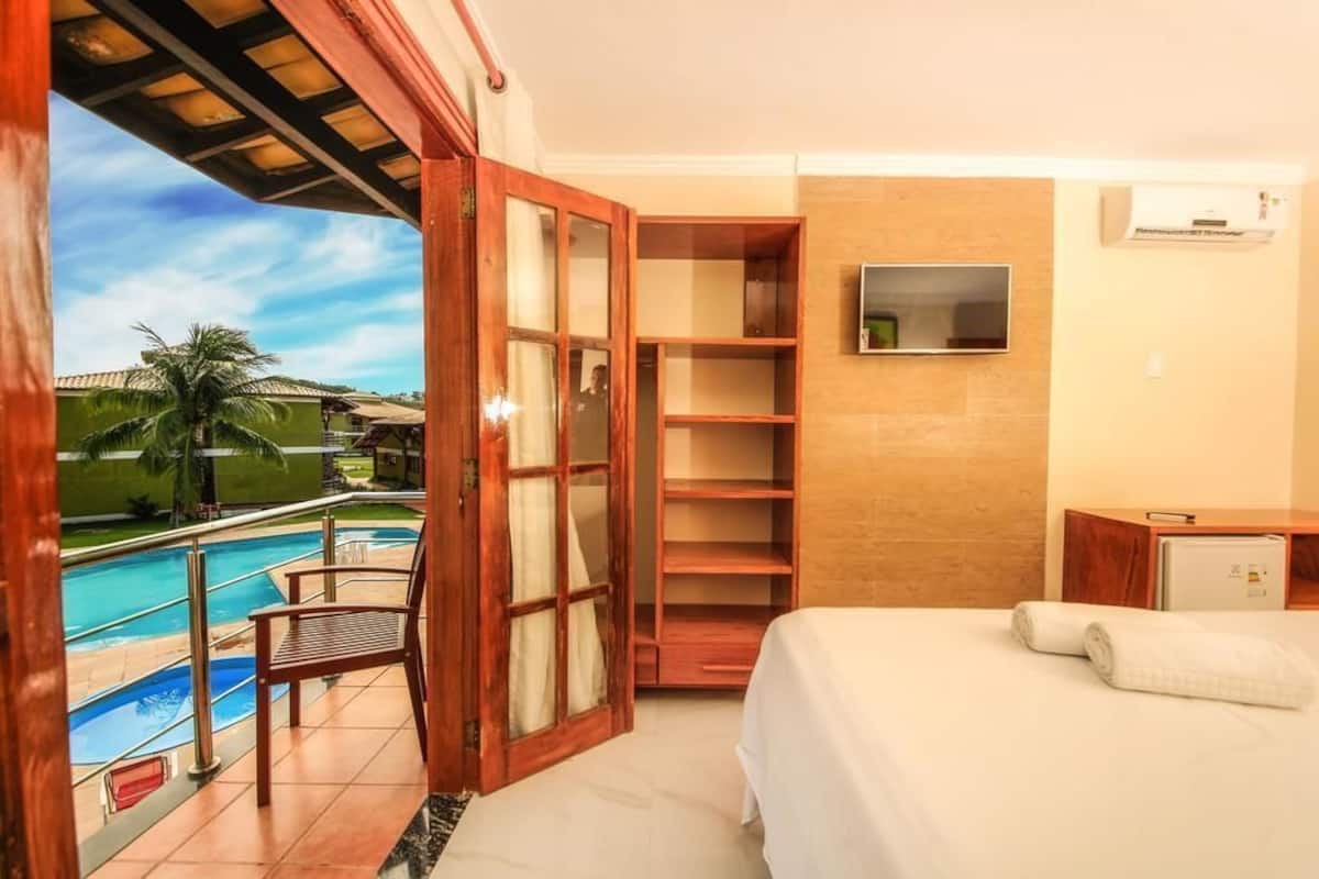 Ein Hotelzimmer mit Bett, Fernseher, einer Holzauslage, Blick auf den Pool und einem Balkon.