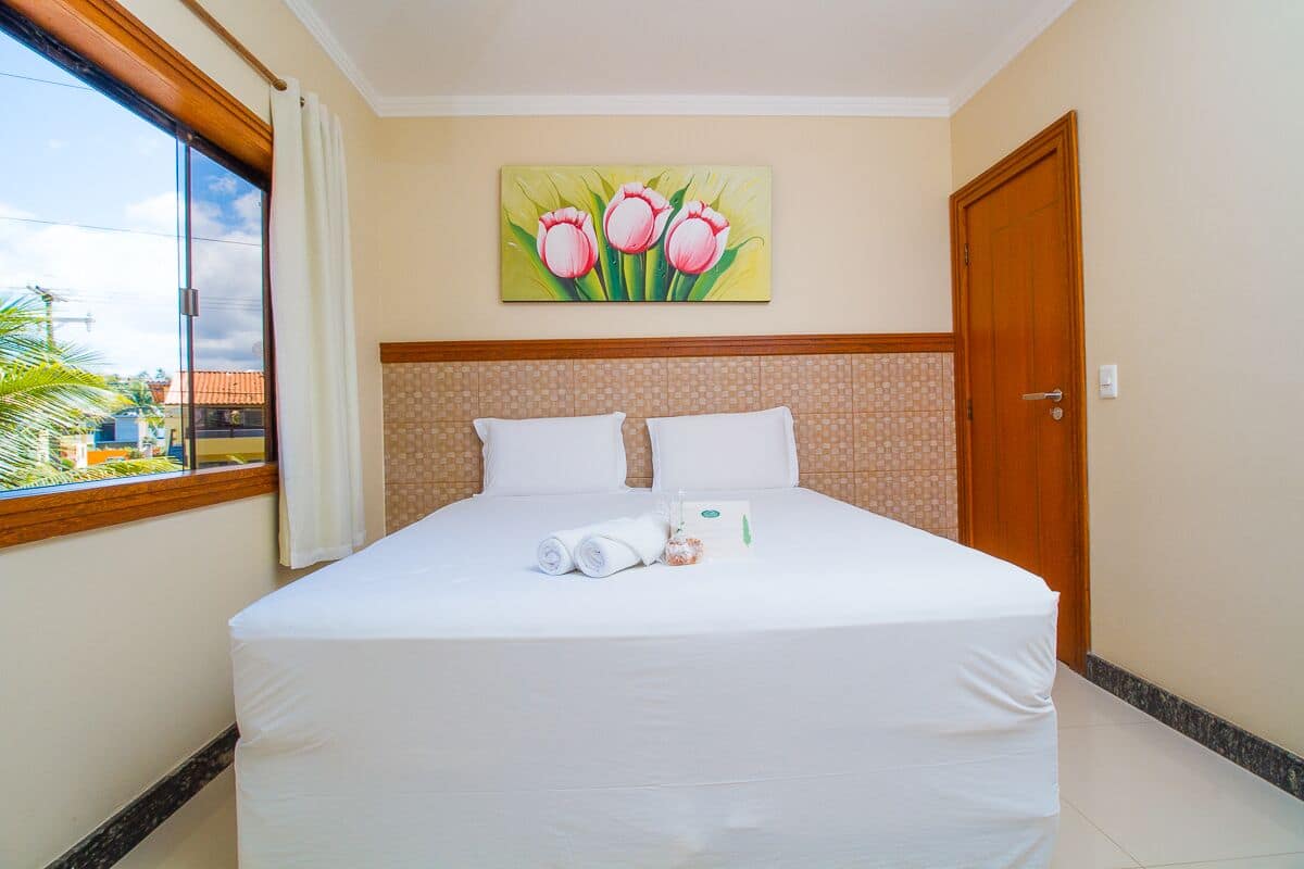 Ein ordentlich bezogenes Bett mit weißen Leinen und Kissen, ein Gemälde von rosa Tulpen an der Wand und eine Holztür zur Rechten.