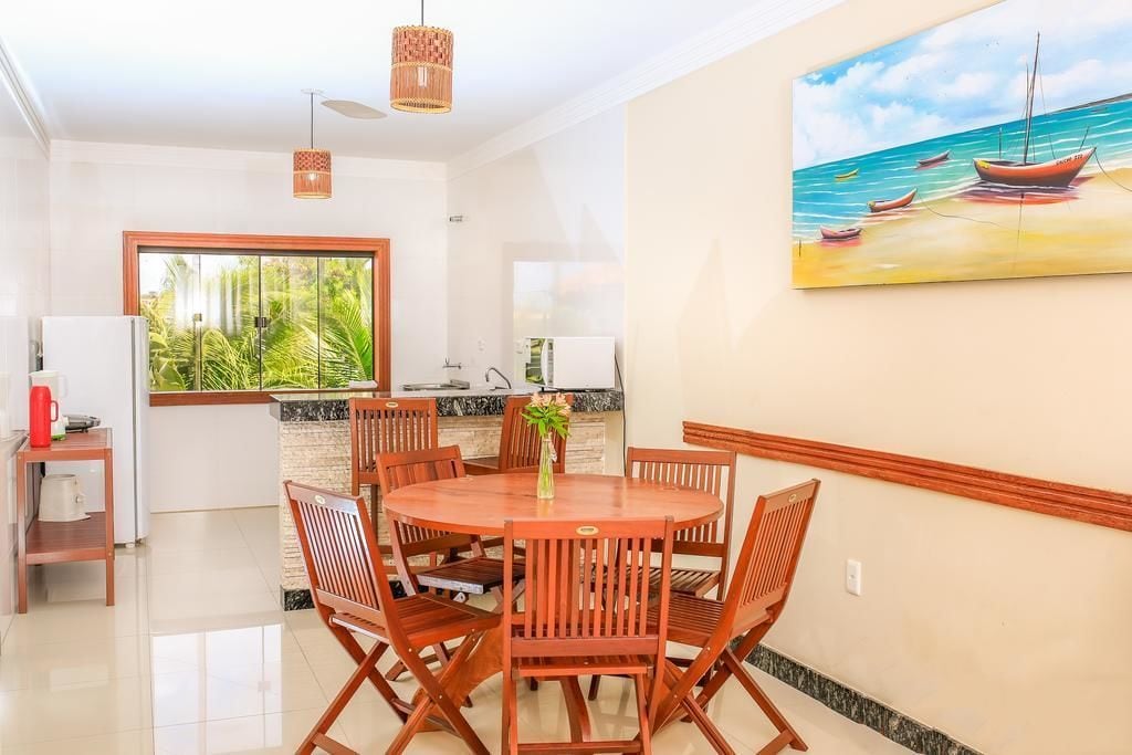 A dining area with a round wooden table and chairs, a painting of a beach scene, and a view of greenery through a large window.