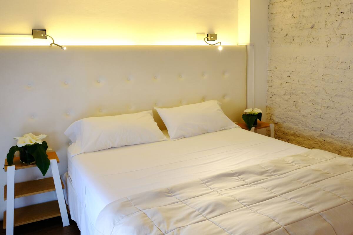 Una cama bien hecha con sábanas y almohadas blancas, una mesita de noche de madera con un florero, y un muro de ladrillo al fondo.