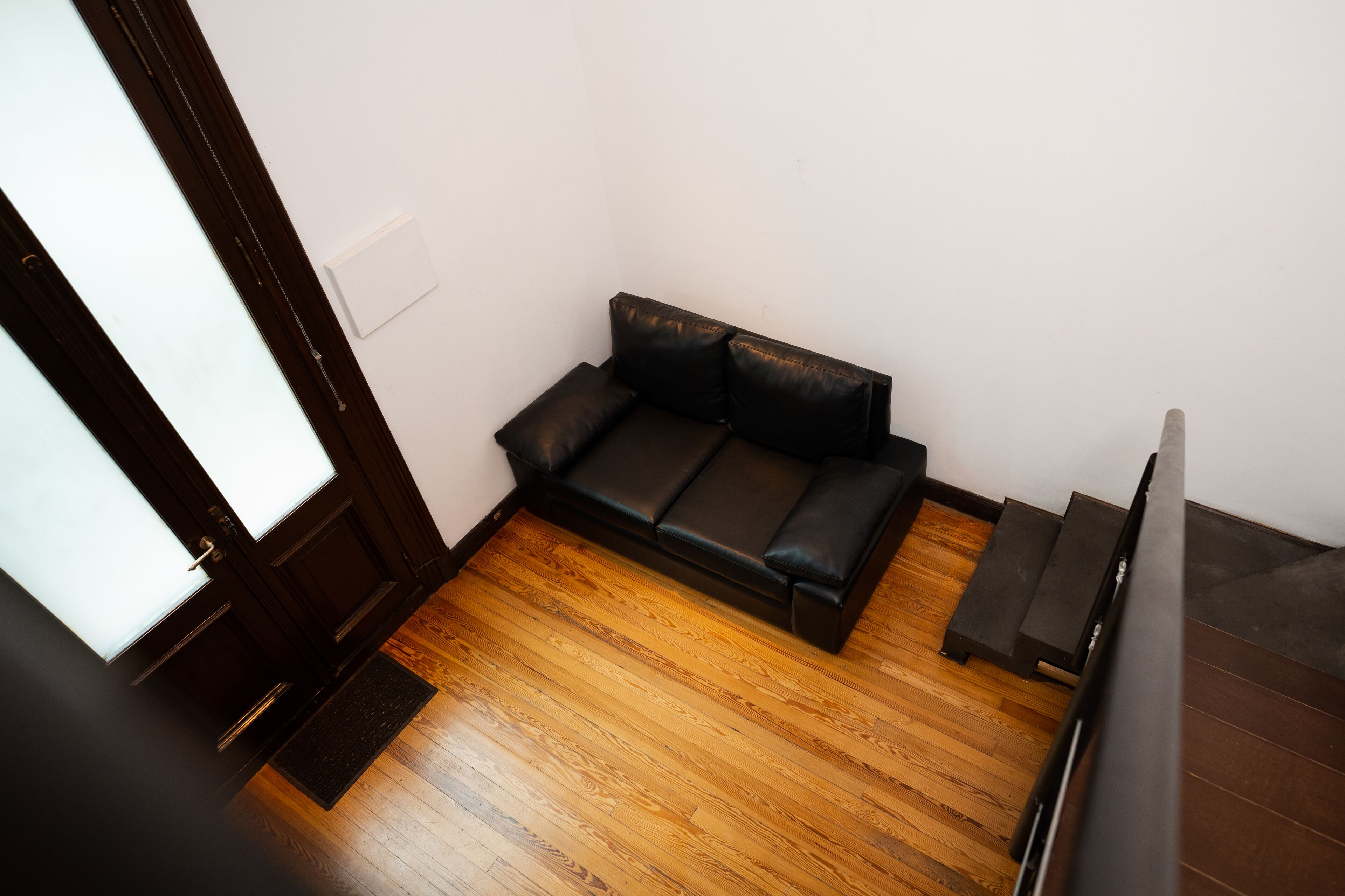 A small, dark-colored sofa in a corner of a room with wooden flooring and a white wall.