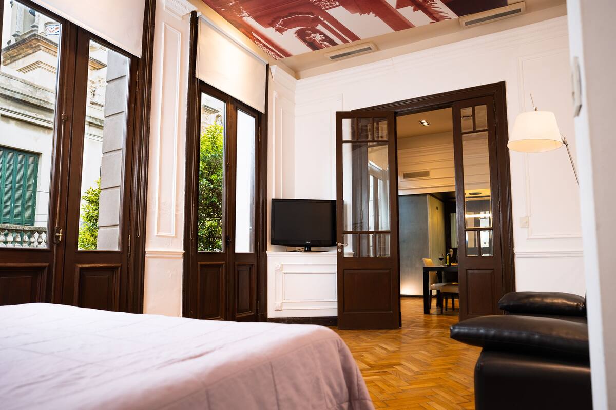 Habitación de hotel con cama, televisión, zona de comedor y vista a la calle.
