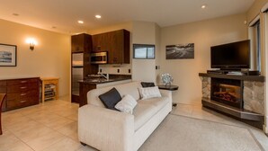 A modern living room with a beige sofa, a flat-screen TV, a fireplace, and a kitchen area with wooden cabinets and a microwave. - The Lagoon Suite at The Estuary Inn (Nanoose Bay)