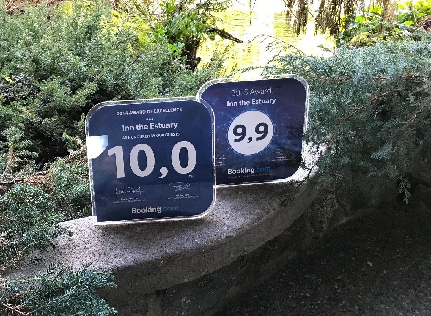 Two awards plaques for Inn the Estuary, one with a 10.0/10 rating and another with a 9.9 rating, placed on a stone ledge surrounded by greenery.