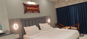Quarto de hotel com uma cama grande, uma escrivaninha, uma cadeira, uma televisão presa à parede e uma placa decorativa sobre a cama.