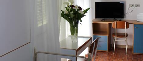 A hotel room with a bed, a desk with a television, a chair, and a window with sheer curtains.