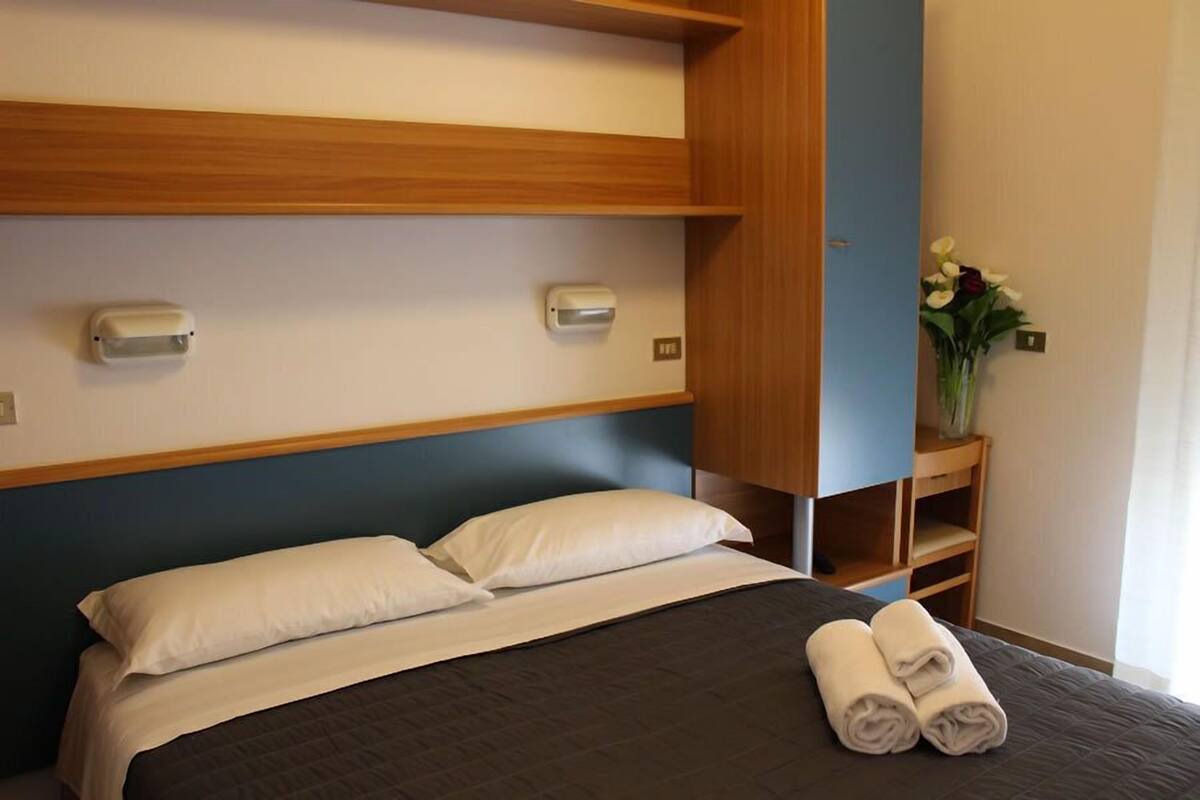 A hotel room with a bed, two wall-mounted light switches, a wooden shelf, and a vase with flowers.