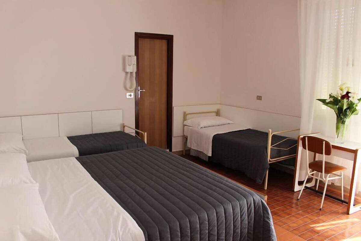 A hotel room with two single beds, a desk with a chair, a telephone, and a window with curtains.