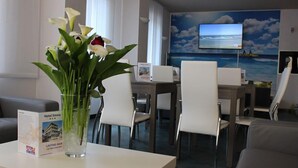 Un vestíbulo de hotel con mostrador de recepción, sillas blancas, zona de comedor y una gran pantalla de televisión mostrando una escena de playa.