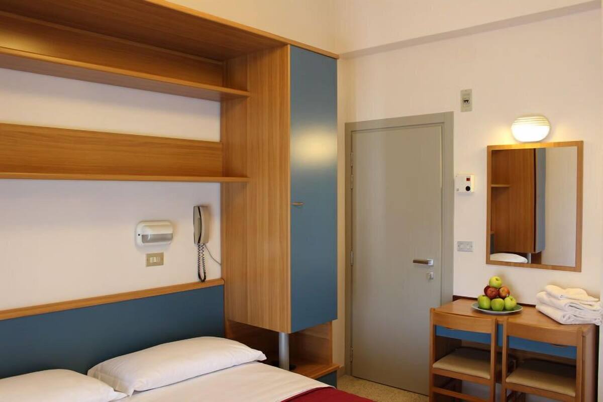A hotel room with a bed, a wooden wardrobe, a desk with a mirror, a telephone, and a bowl of fruit.