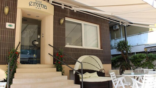 Entrada de un hotel con un letrero que dice "hotel Emma", una escalera que conduce a la entrada, zona de estar al aire libre con sillas y mesas blancas, y un área de descanso cubierta con techo curvo.