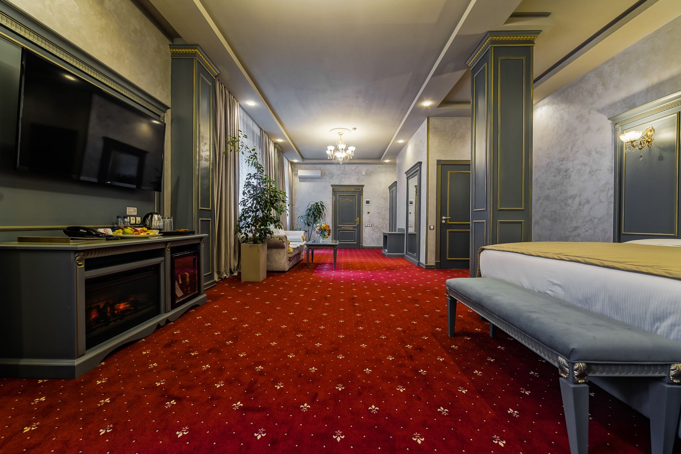 A hotel room with a red carpet, a large bed, a bench, a TV, and a fireplace.